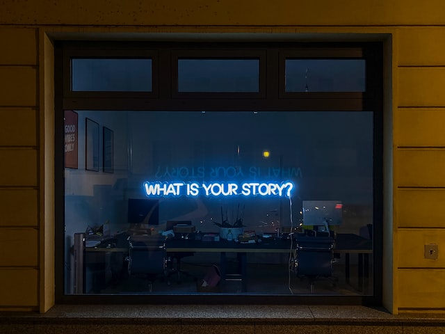 A neon sign on a dark window reads: What Is Your Story?